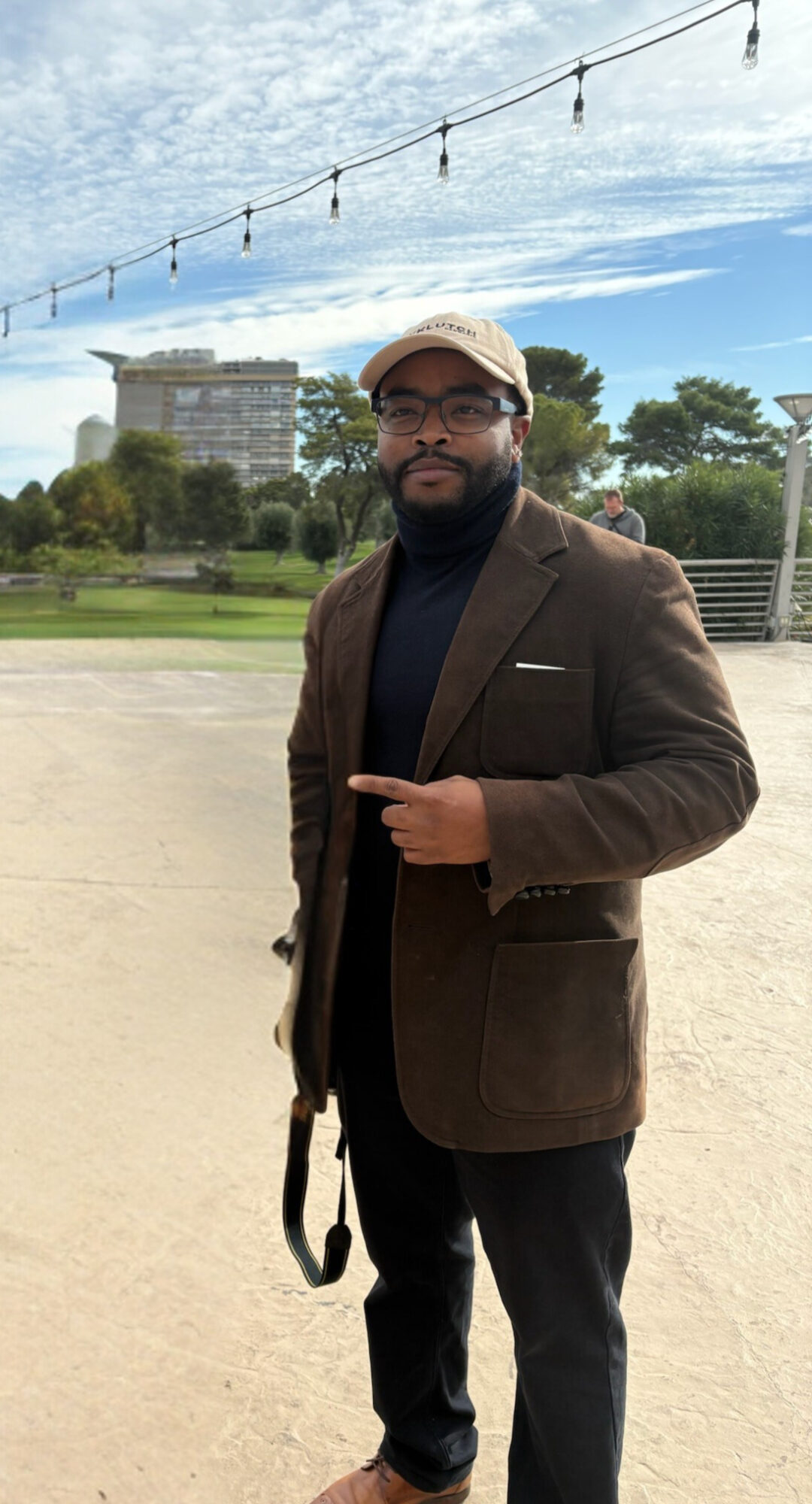 Man in brown blazer and black turtleneck standing outdoors with trees and buildings in background.