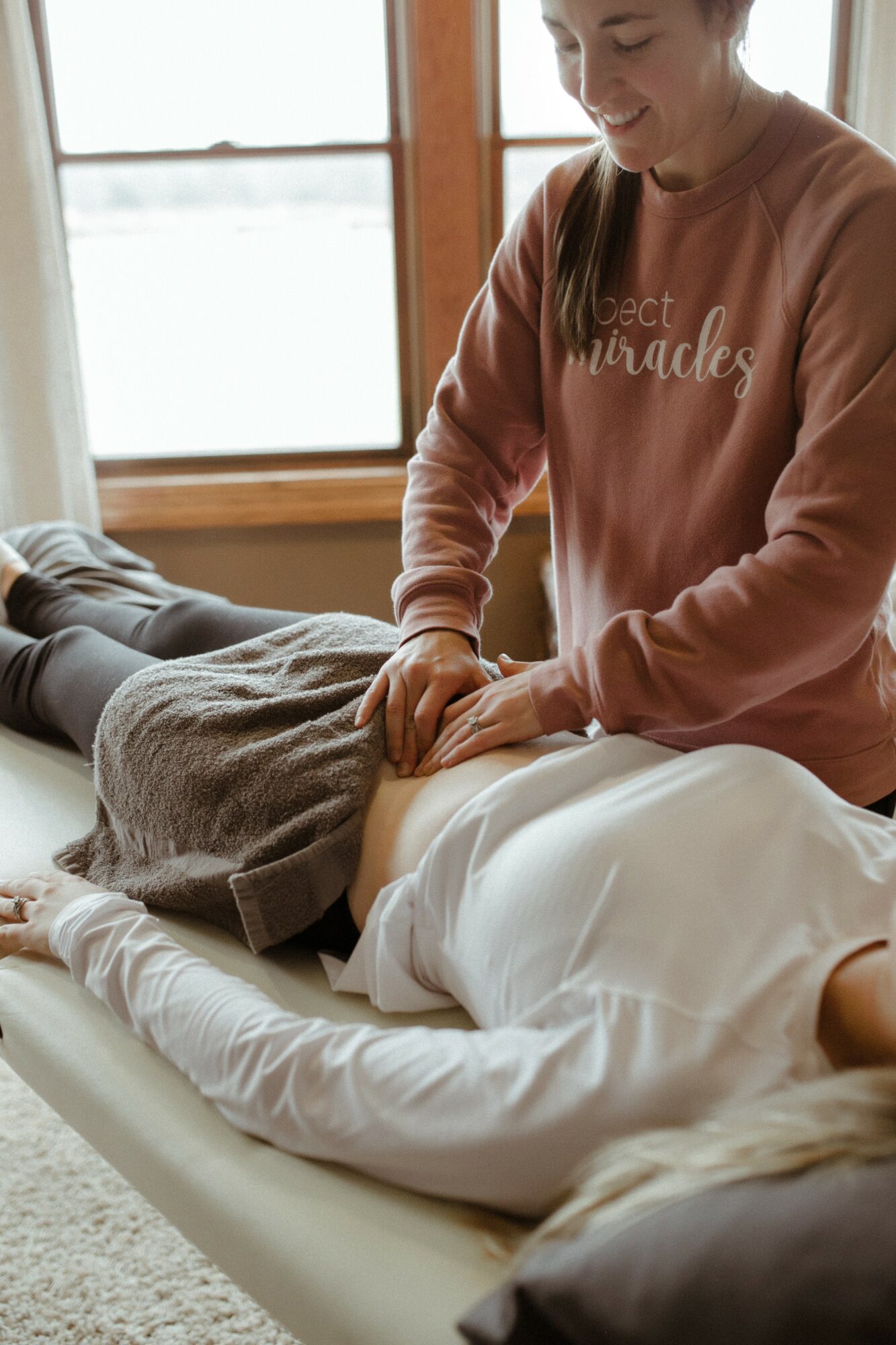 Person receiving a massage on their abdomen from a therapist in a room with large windows, smiling, wearing a pink sweatshirt.