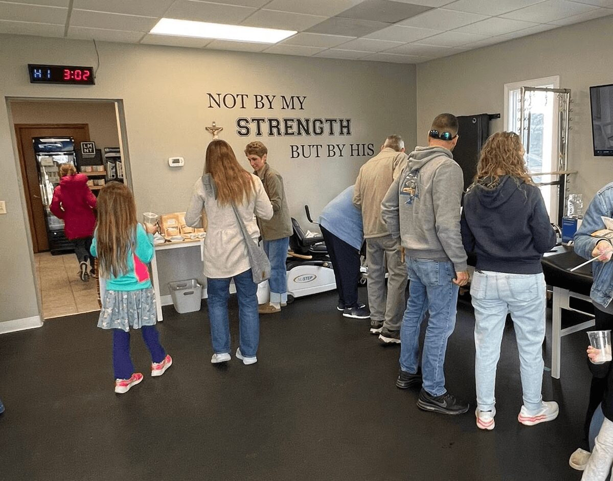 People standing in line inside a room with a wall that has a motivational quote, some children, and a digital clock showing 3:02.