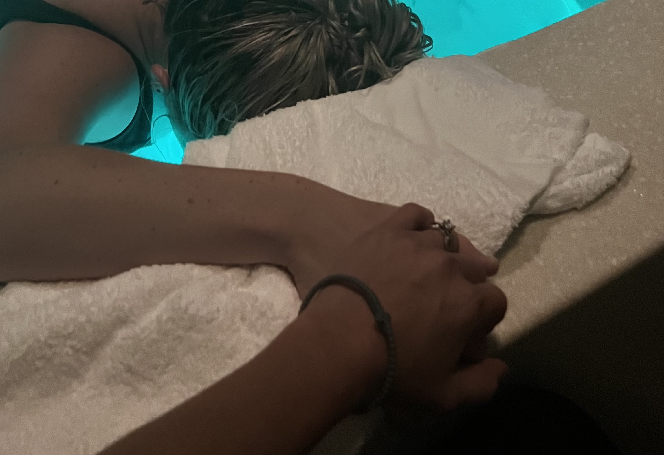 Person lying face down with head on a towel, hand holding another person's hand, in a pool or spa setting.