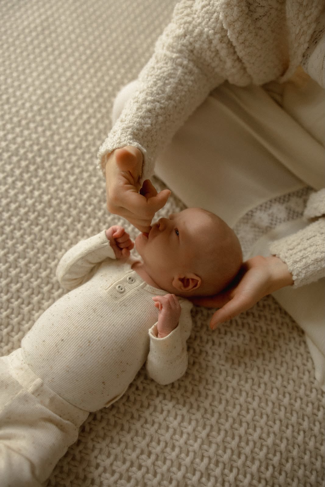 Adult holding a baby on a textured surface, with the baby reaching for the adult's hand, both lying down.