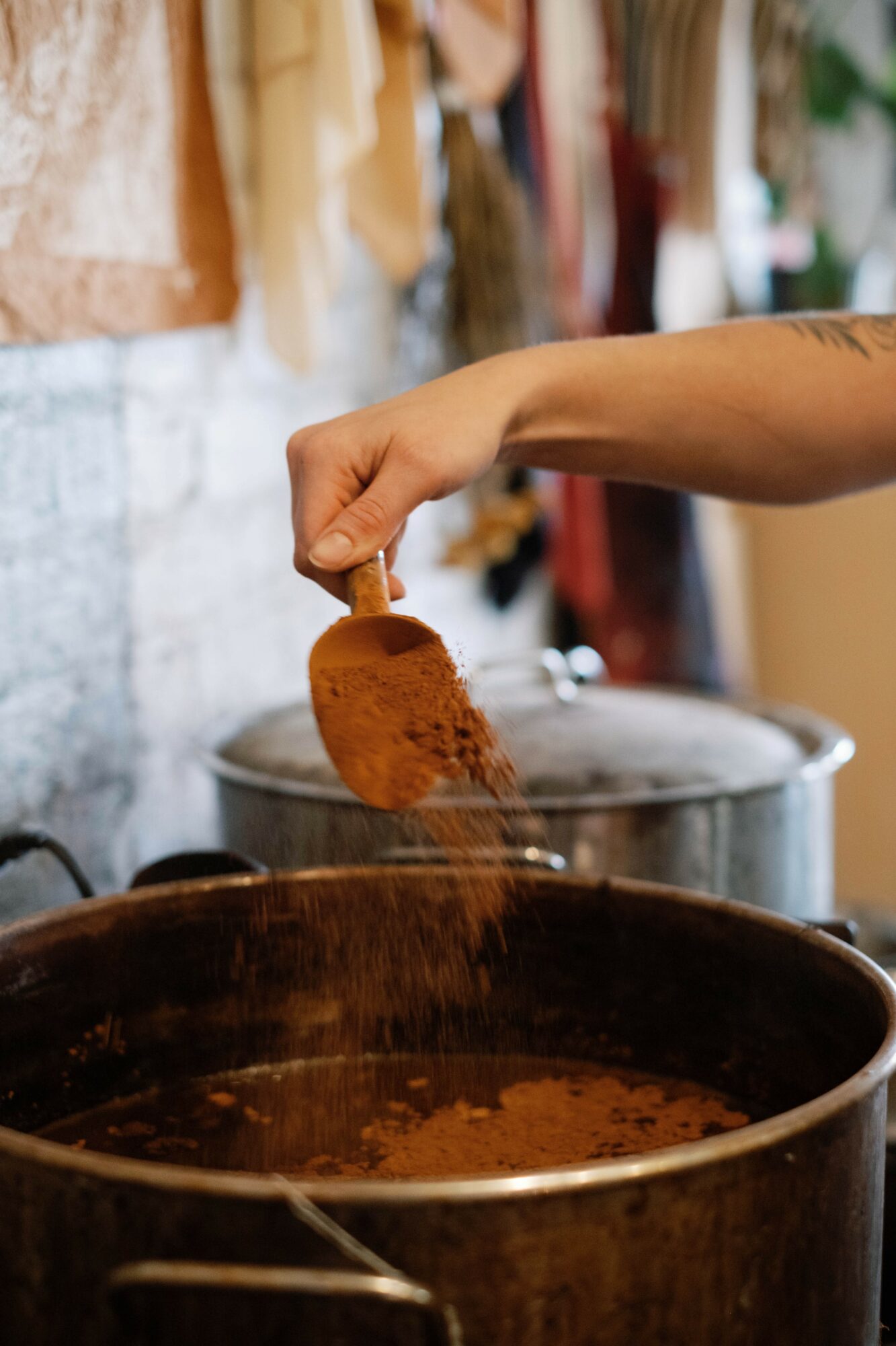 Person's hand holds a spoon with brown powder over a pot of food, in a kitchen setting.
