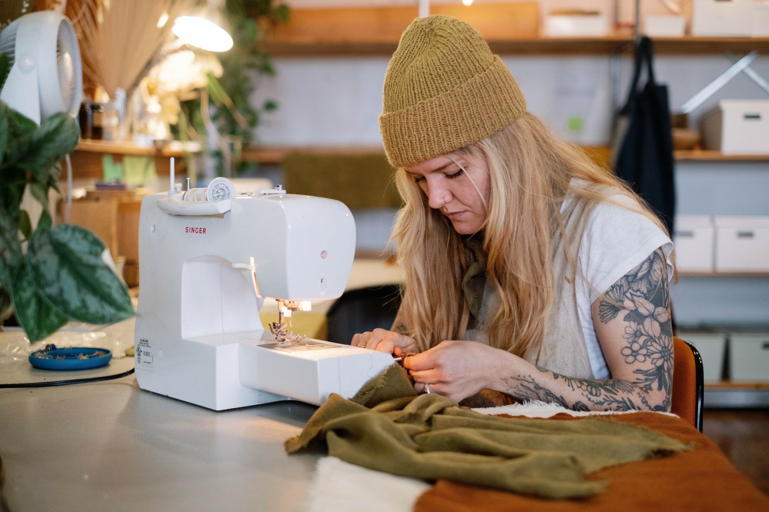 Woman sewing with a sewing machine at a table, wearing a yellow beanie and glasses, with fabric nearby.