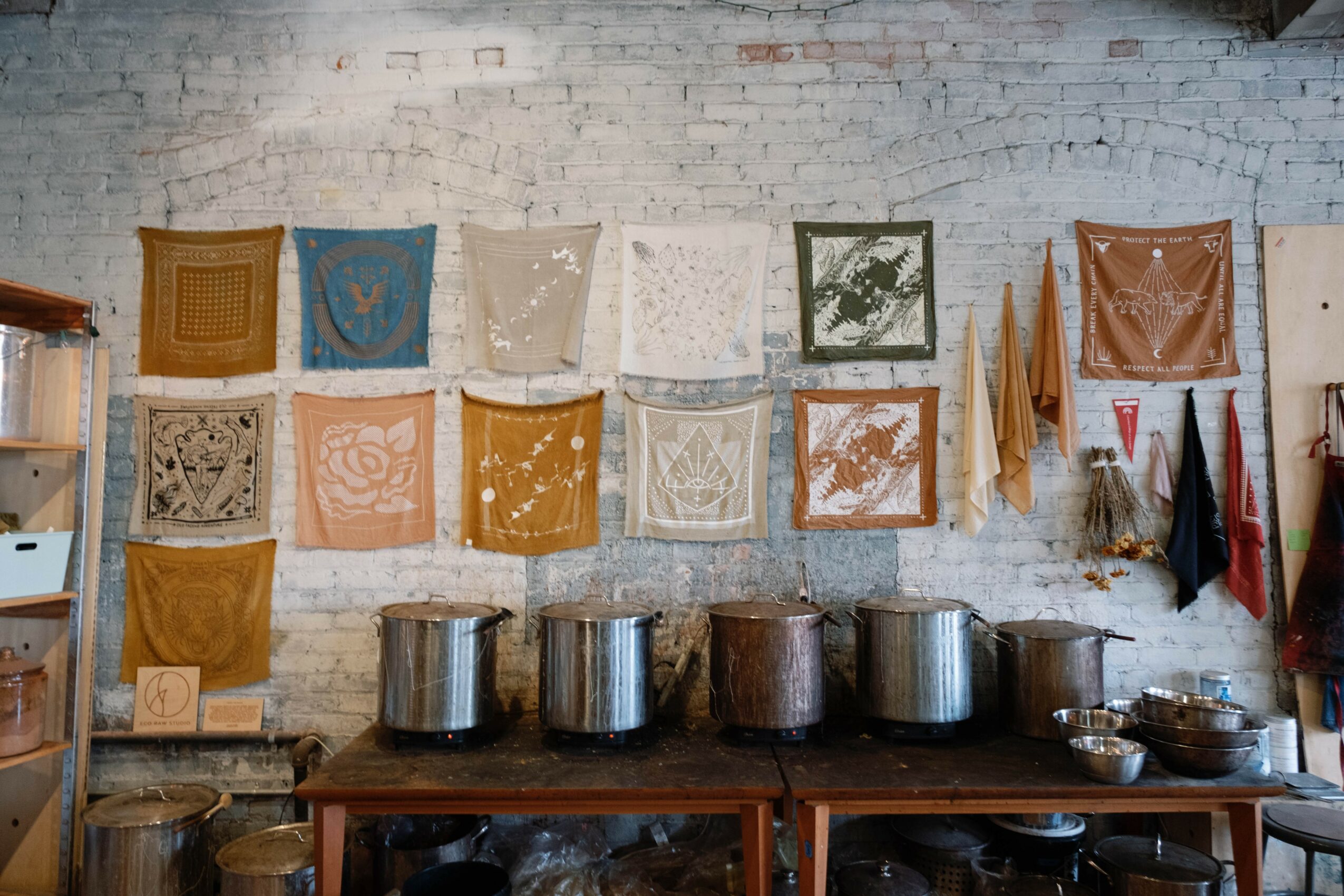 Display of colorful fabric pieces hanging on a white brick wall above a wooden table with large metal pots.
