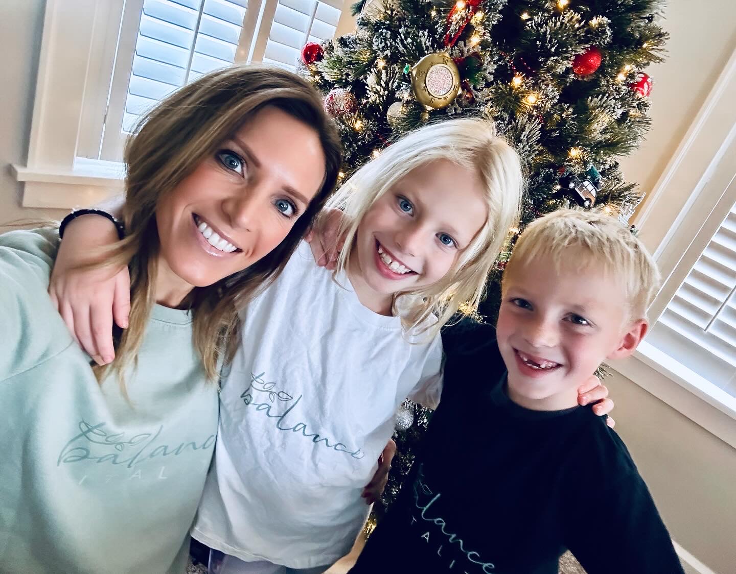 Three smiling people, two children and an adult, standing in front of a decorated Christmas tree indoors.
