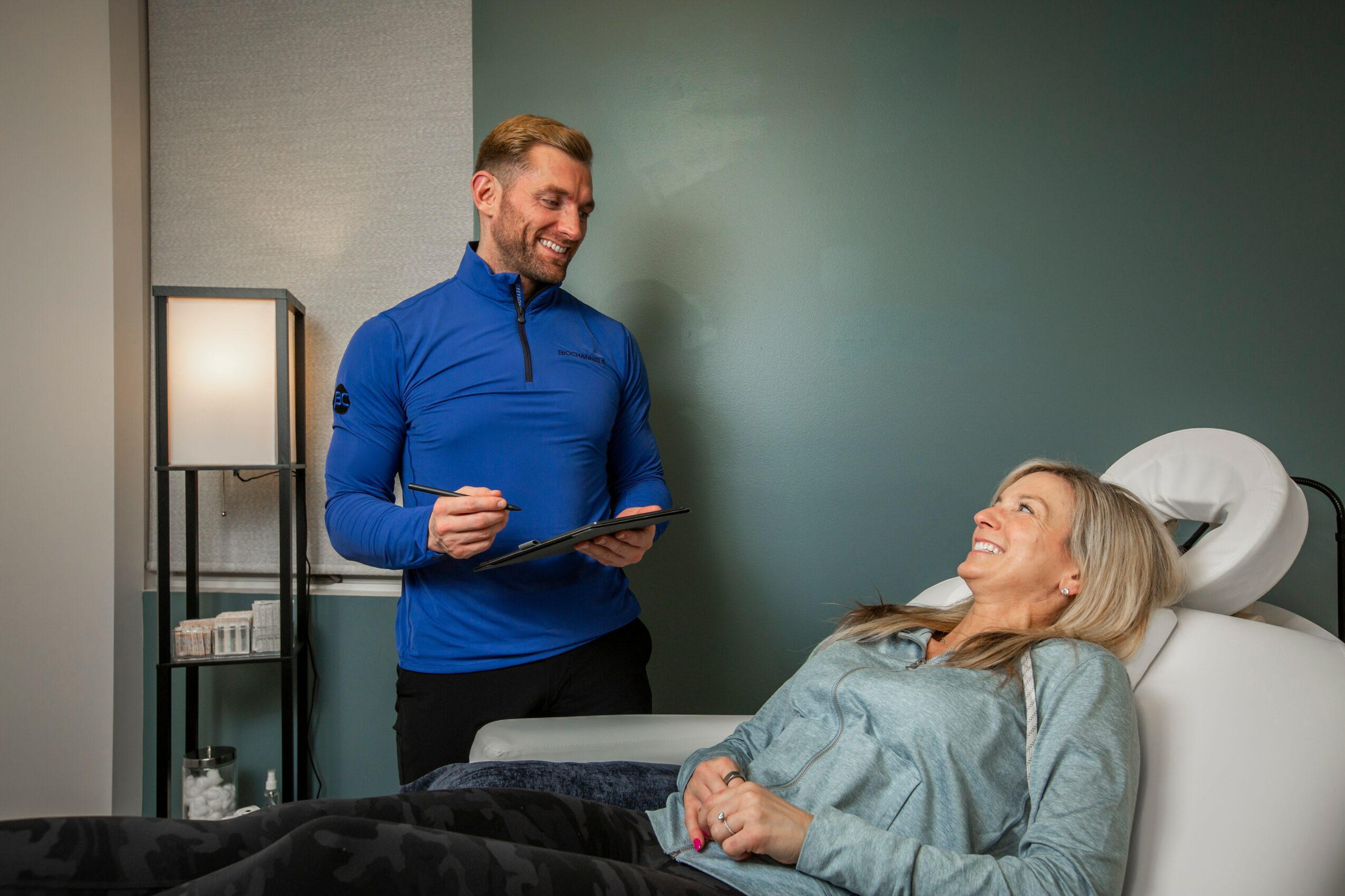 A man in a blue jacket smiling and holding a clipboard stands next to a woman lying in a hospital bed, smiling back.