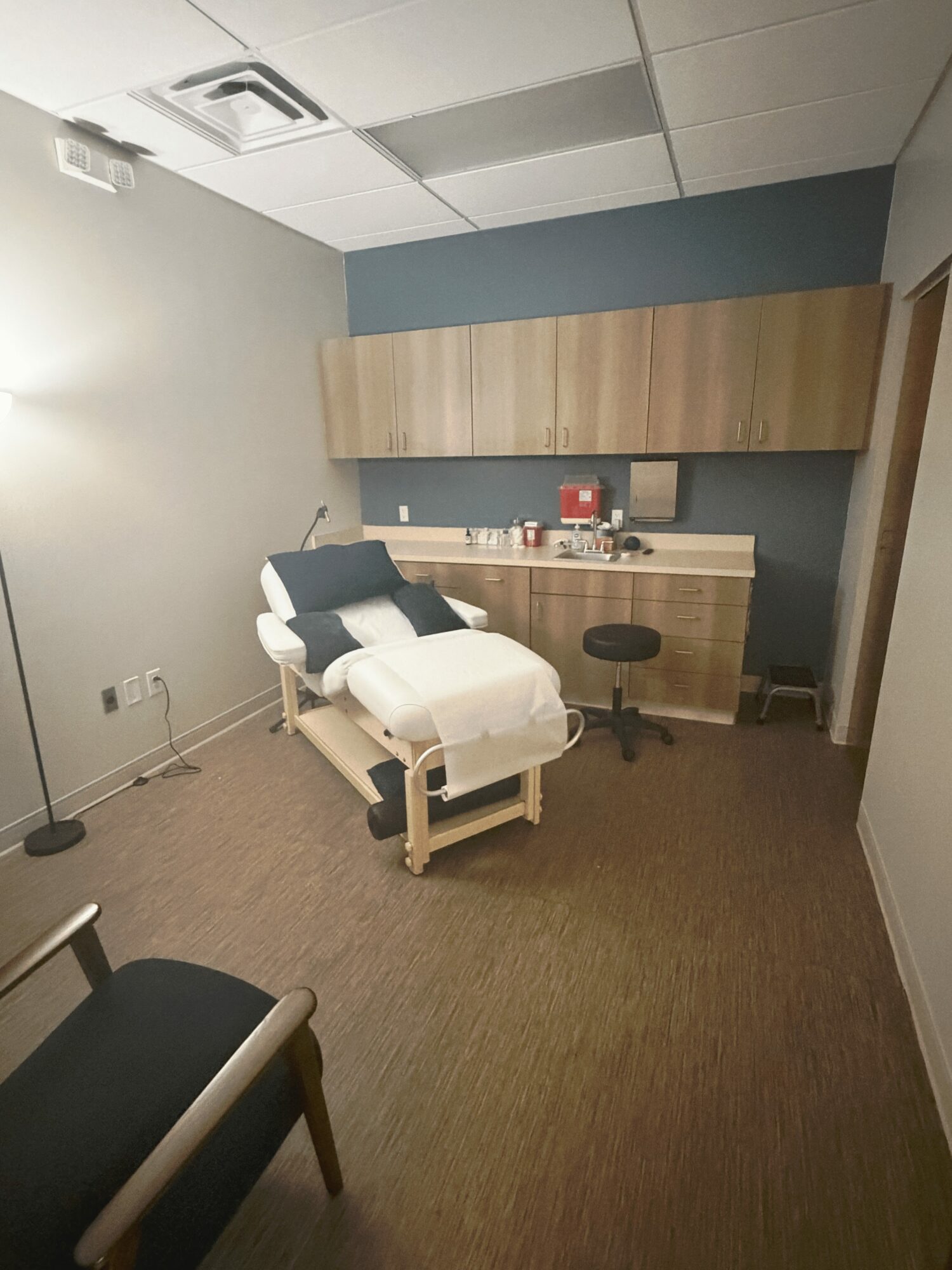 Medical examination room with examination table, cabinets, and chairs, illuminated by ceiling lights, beige carpeted floor, and blue wall.