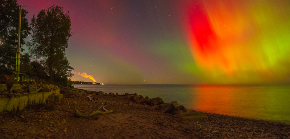 Night sky with colorful aurora borealis over a rocky shoreline, trees, and calm water, with a distant light on the horizon.