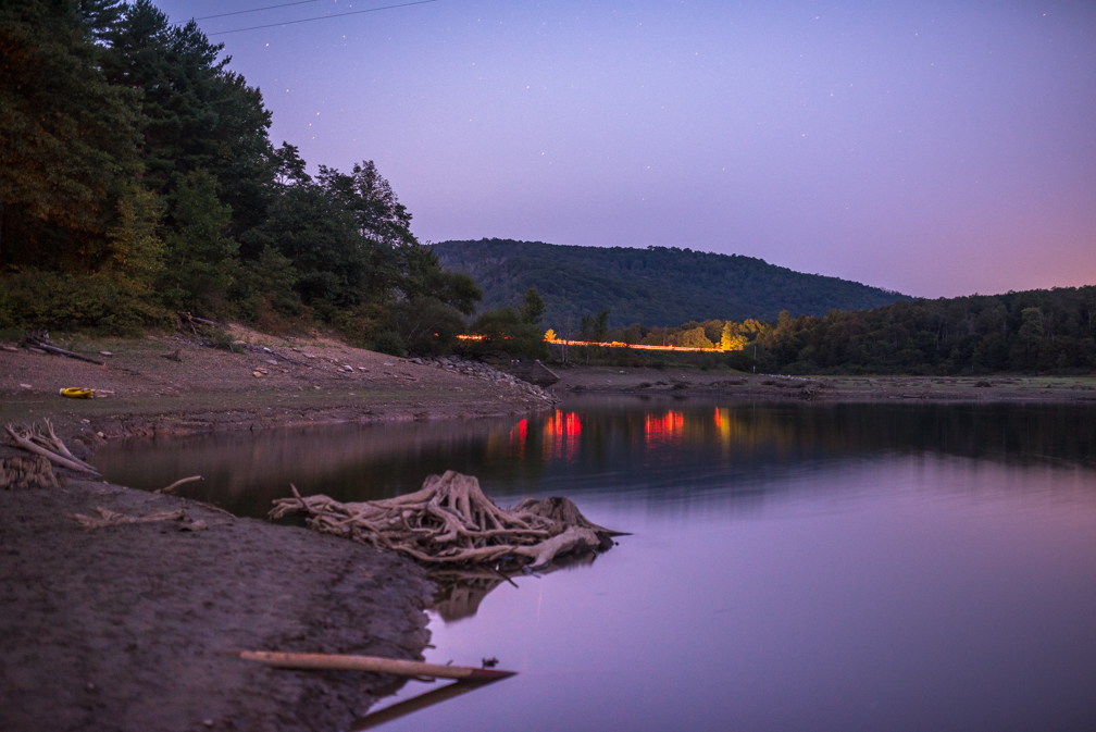 Calm river with trees on the bank and a distant hill under a purple sky, reflecting lights on the water surface.