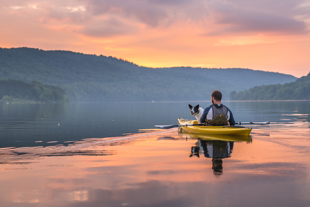 Person kayaking on a calm lake with a dog, surrounded by hills and a colorful sunset sky.