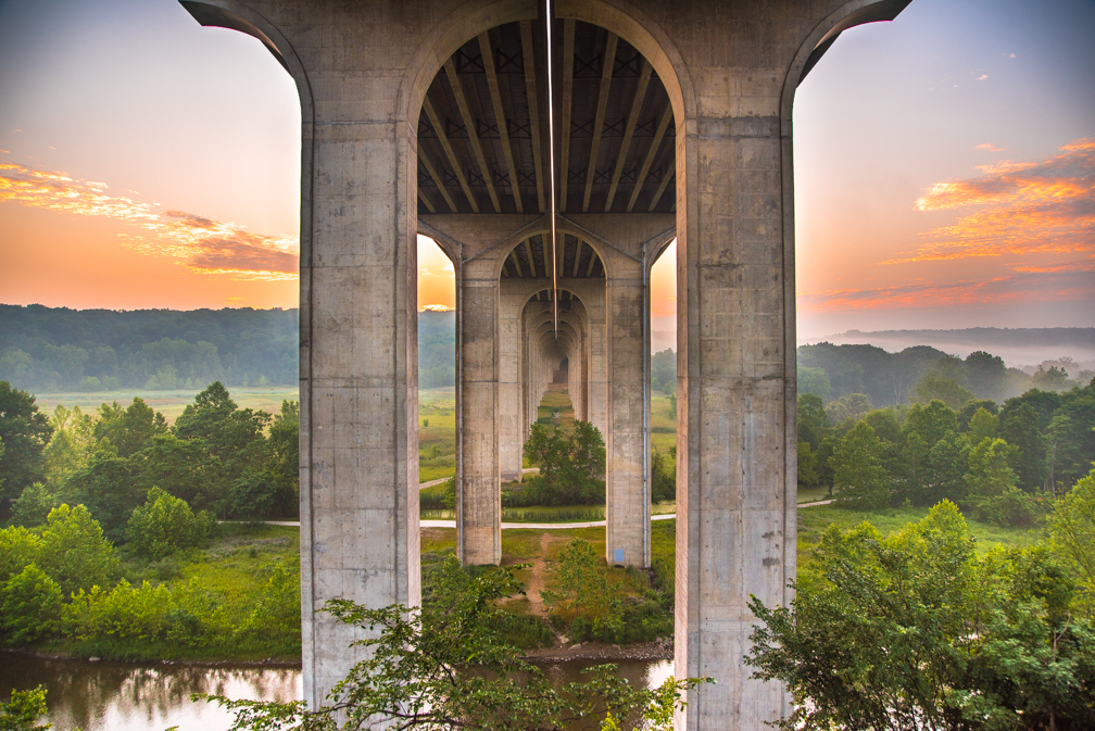 View of a bridge with large concrete pillars over a lush green landscape at sunrise or sunset.