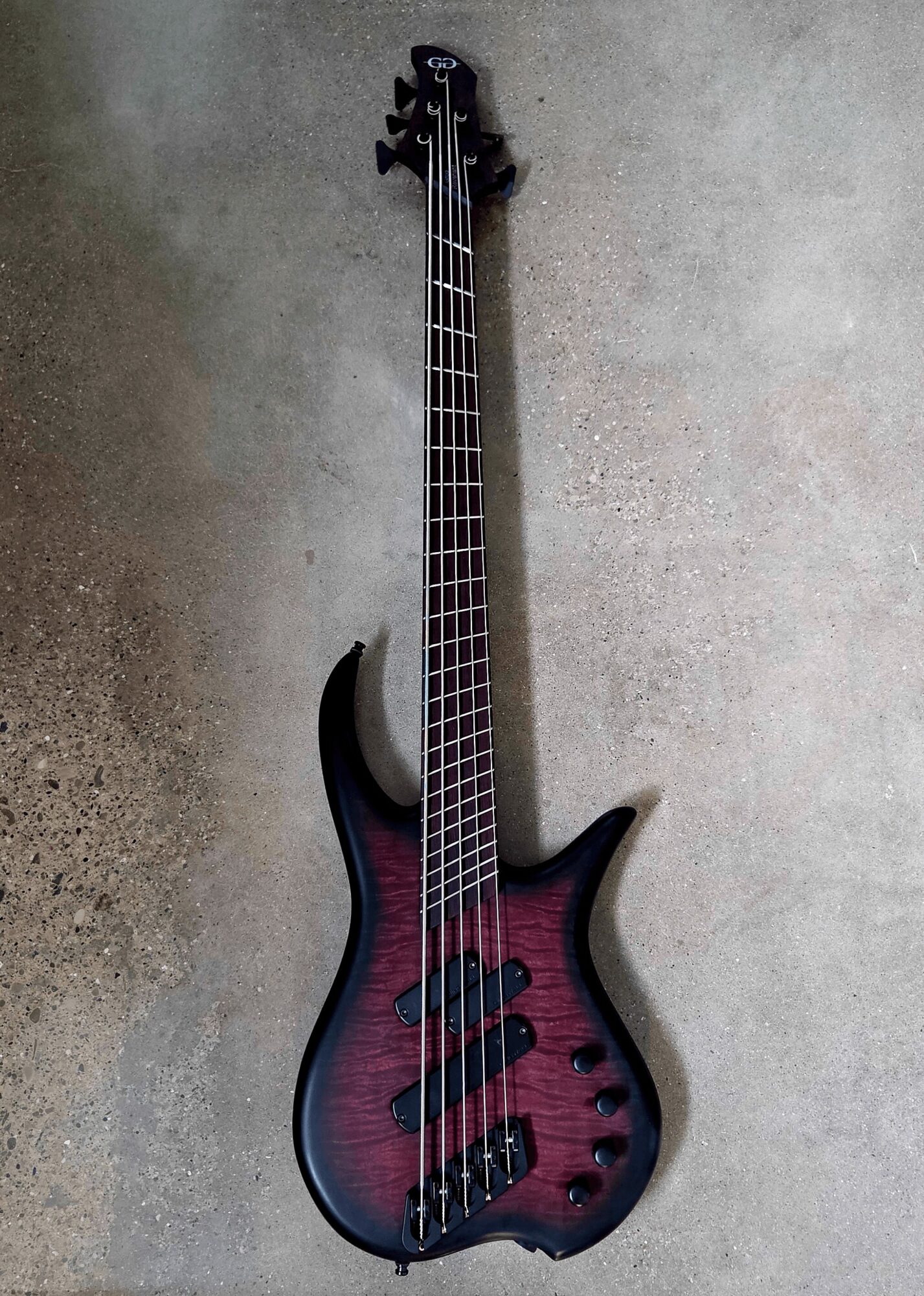 Electric bass guitar with a dark red and black finish, four strings, and a unique body shape, standing upright against a plain wall.