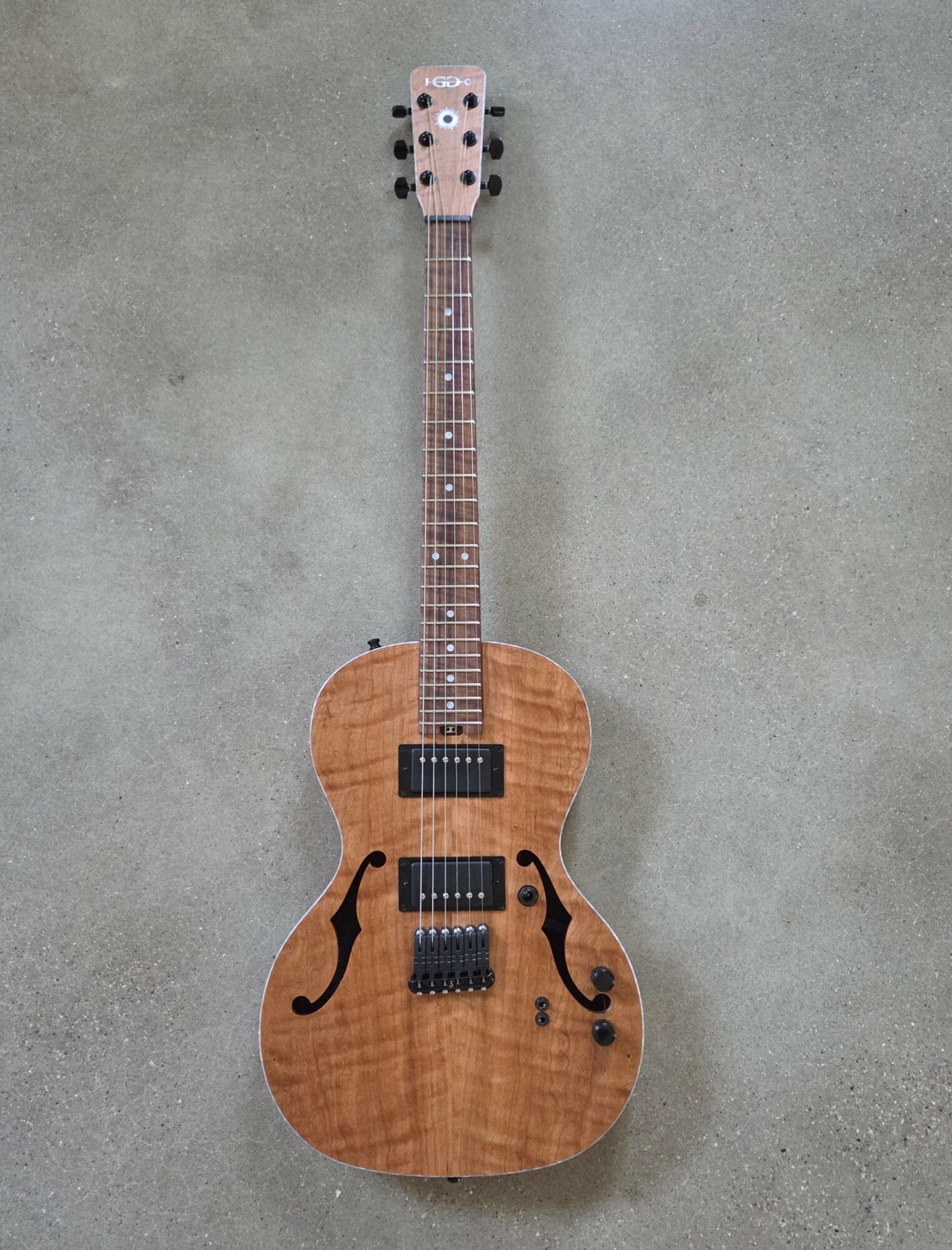 Wooden electric guitar with two pickups, a bridge, and control knobs, lying on a gray surface.