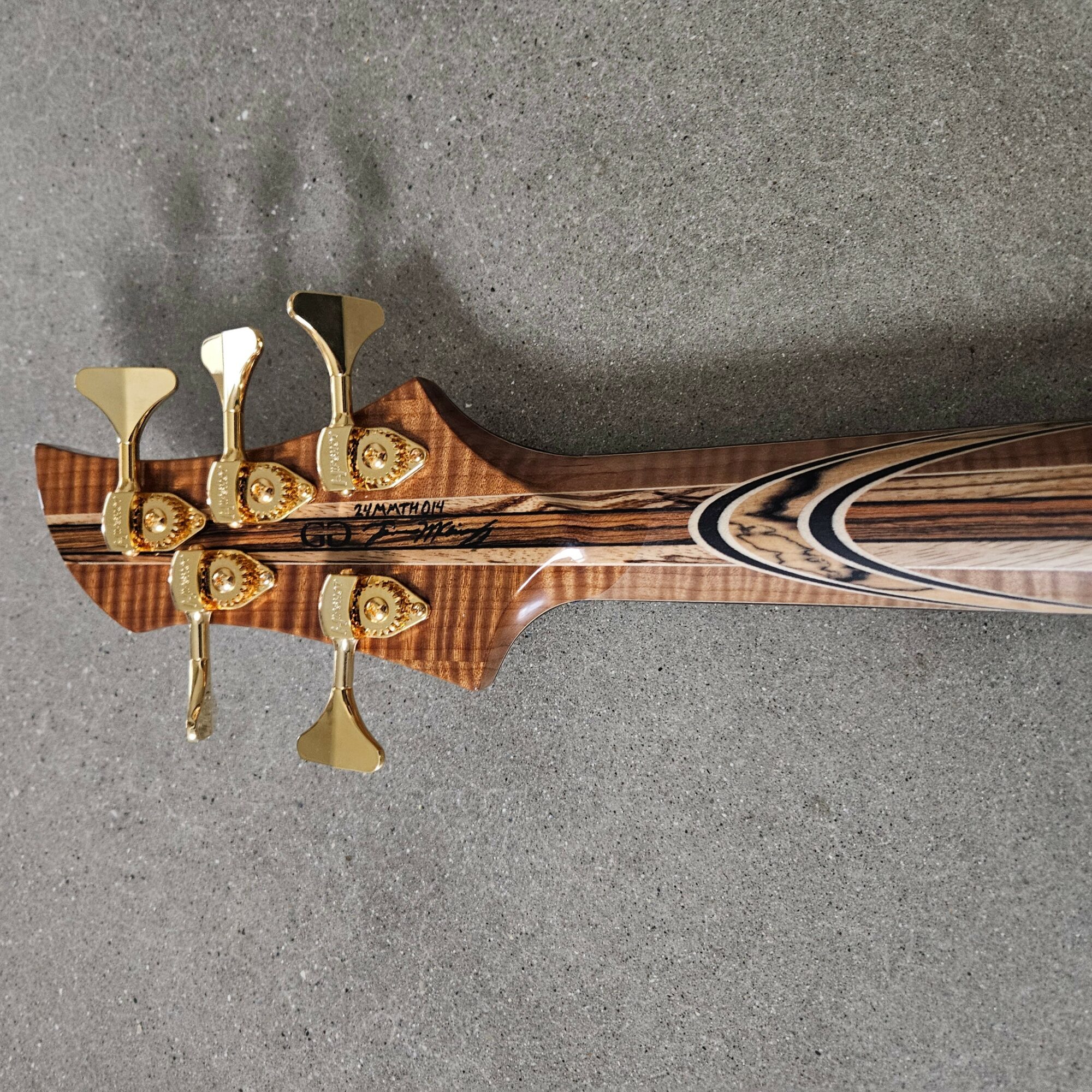 Close-up of a guitar headstock with gold tuning pegs and a wooden neck, on a gray surface.