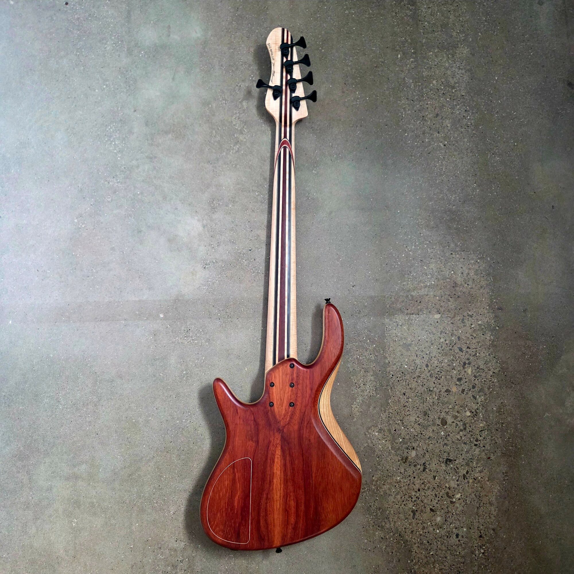 A wooden electric bass guitar with a natural finish, black tuning pegs, and a curved body, hanging vertically against a textured wall.