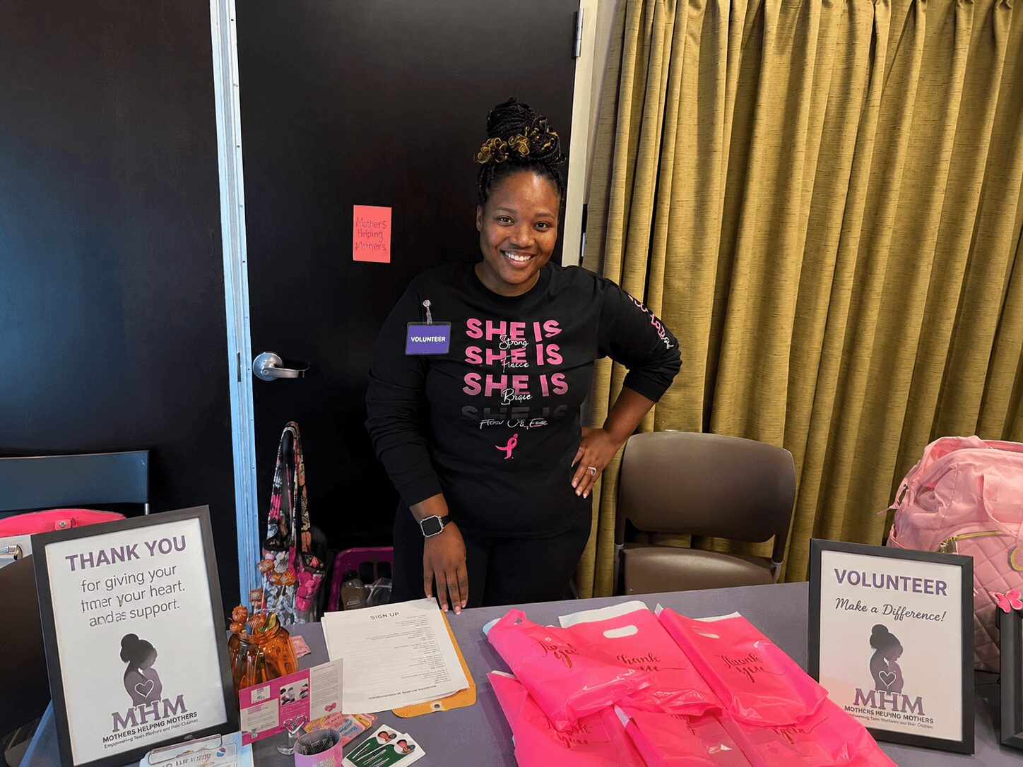 Smiling woman standing behind a table with pink items, surrounded by signs and a curtain, in an indoor setting.