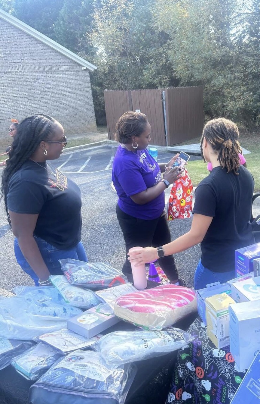 Three women stand outdoors near a table with various packaged items, engaging in conversation, with trees and a fence in the background.