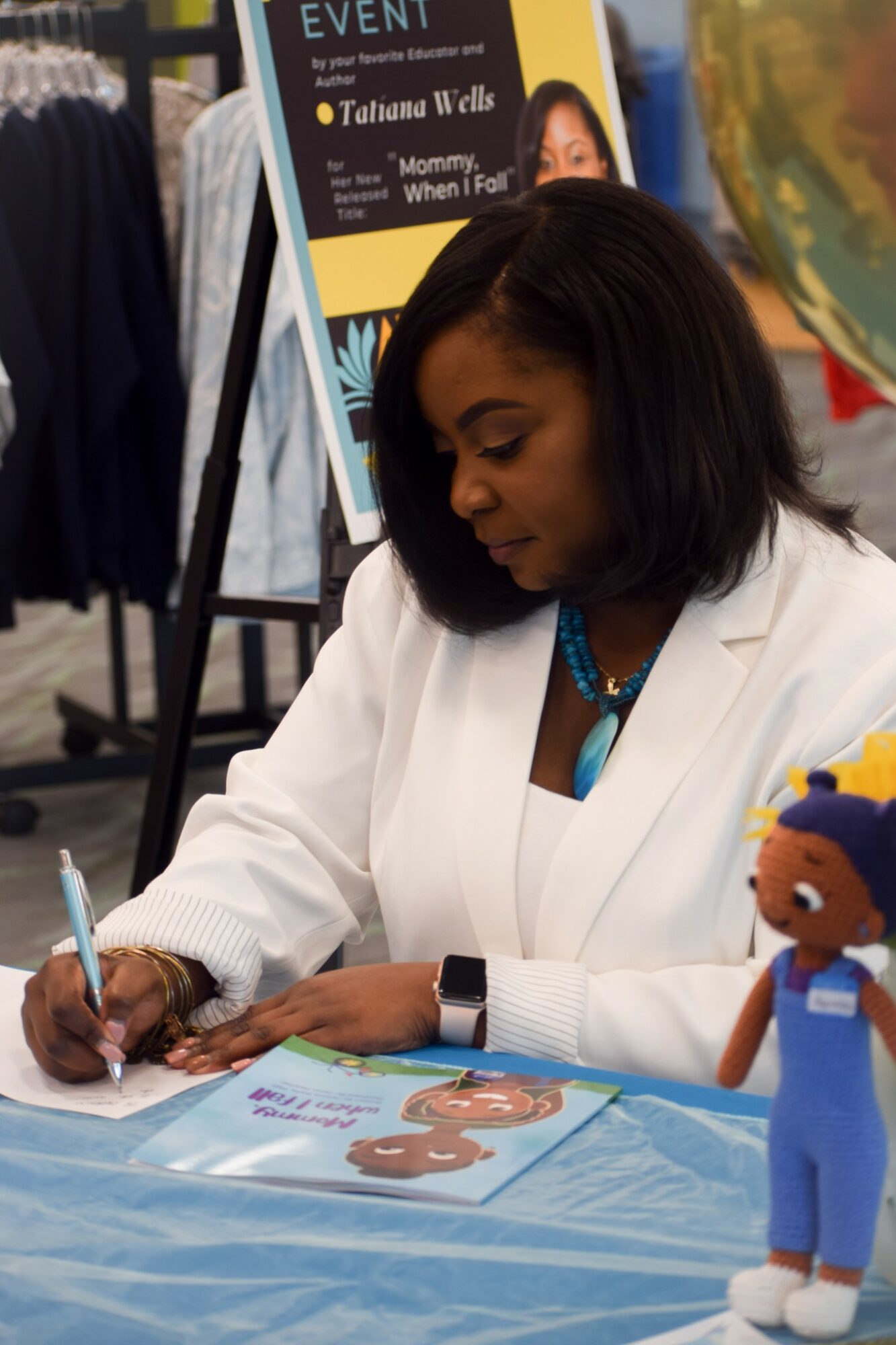 Woman with shoulder-length black hair signing a book at a table, with a colorful poster and a doll nearby.