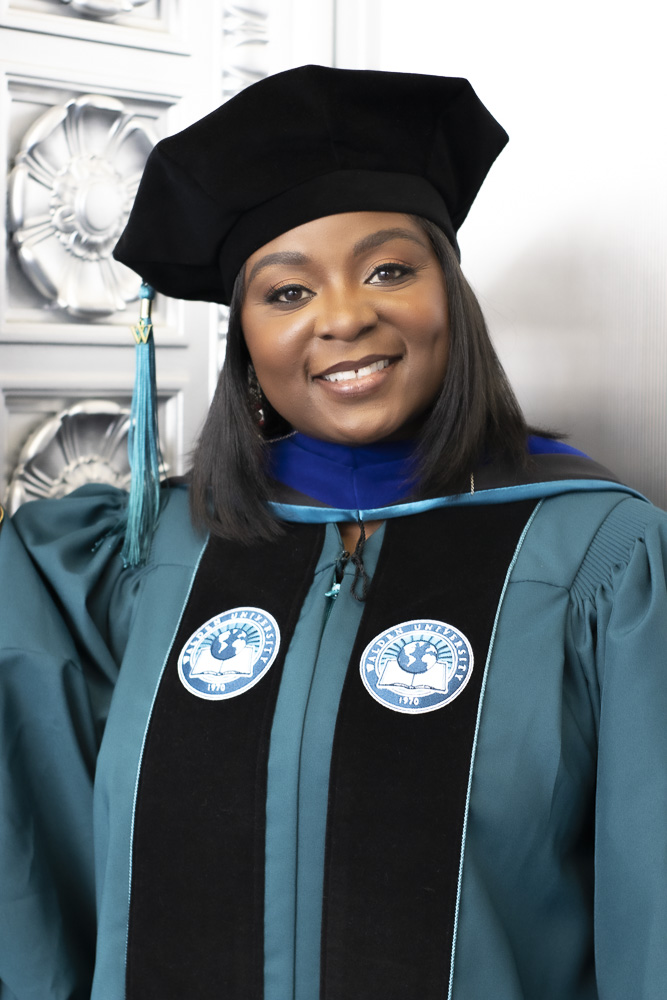Woman in graduation gown and cap smiling, standing indoors with decorative background, wearing teal and black academic regalia.