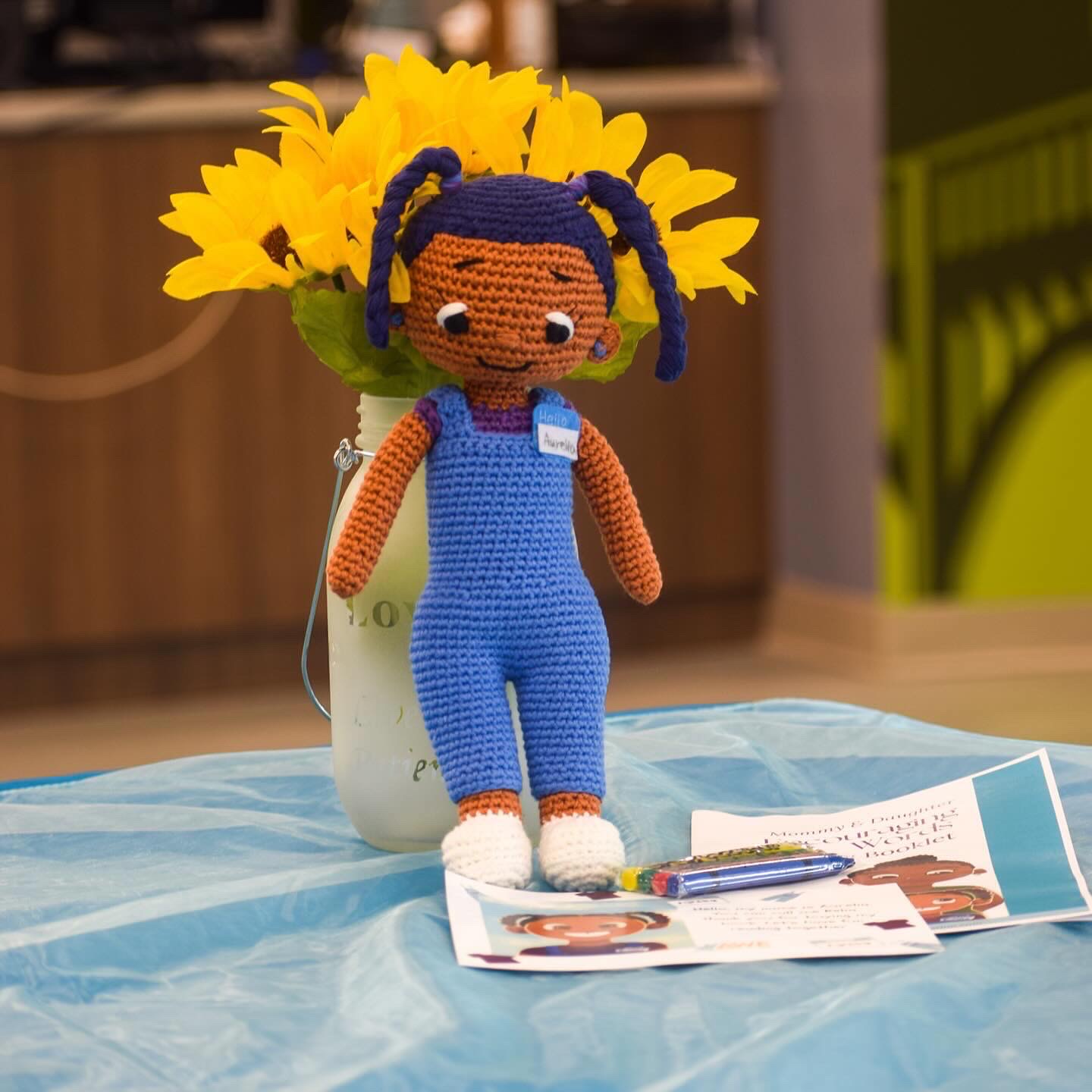 Crocheted doll with blue overalls and pigtails stands in front of a sunflower arrangement, on a table with papers and a pen.