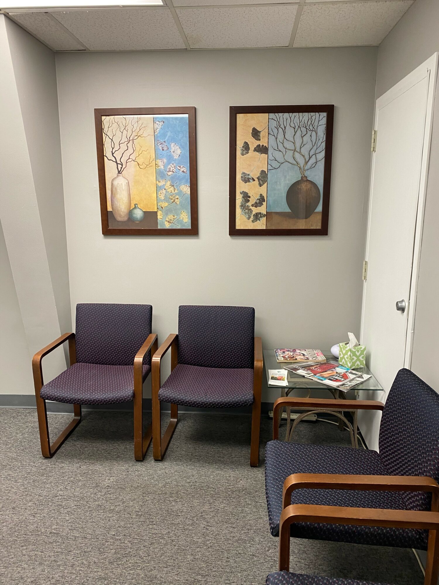 Three chairs and a small table with magazines in a room with two framed paintings of vases and trees on the wall.