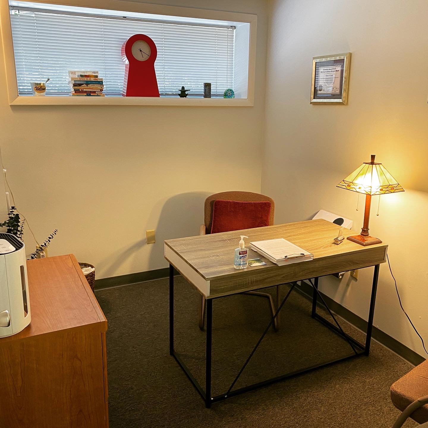 Office corner with desk, chair, lamp, and window with a red clock on the sill.