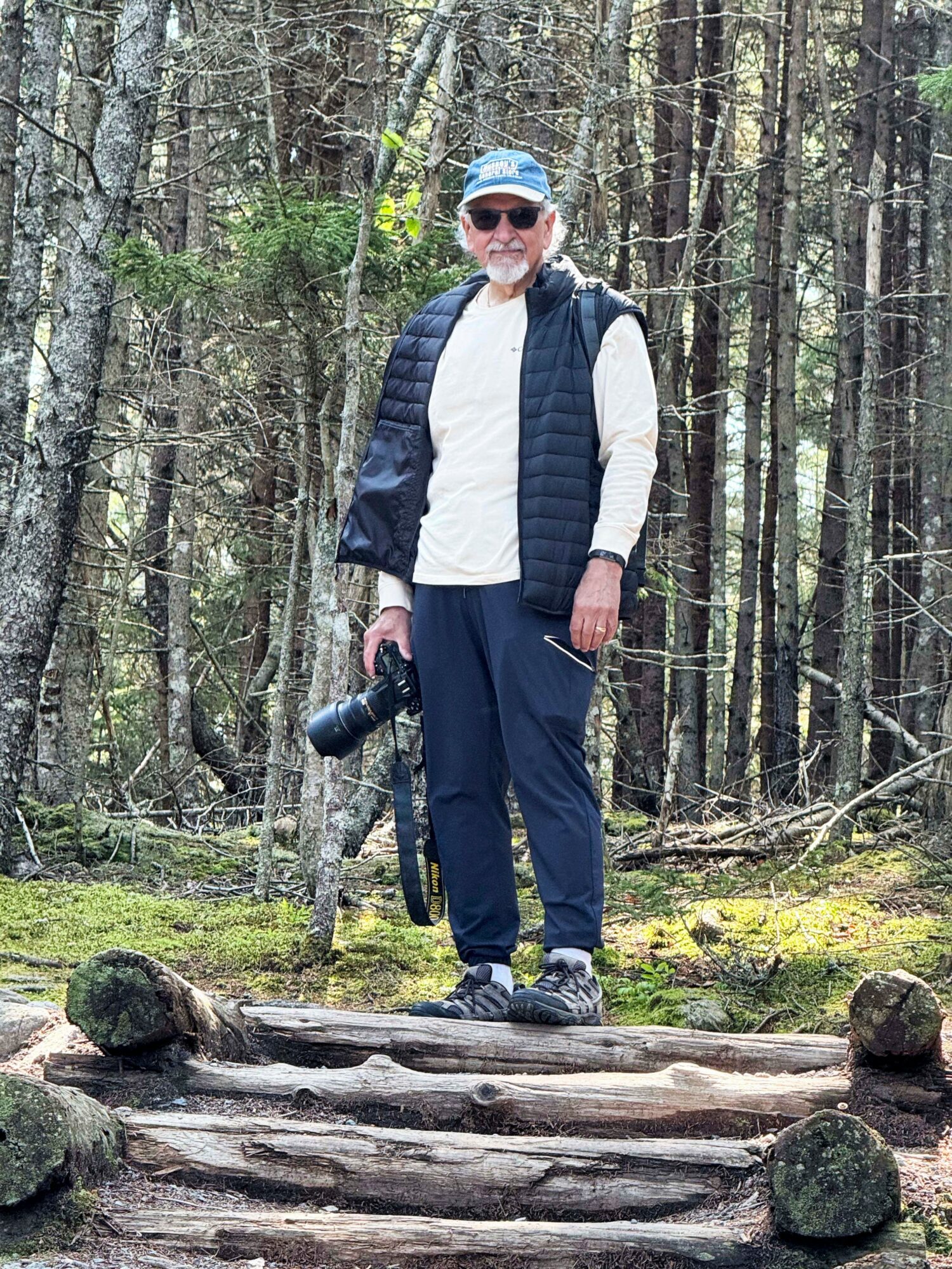Man standing on logs in a forest, holding a camera, wearing sunglasses, a cap, and outdoor clothing.