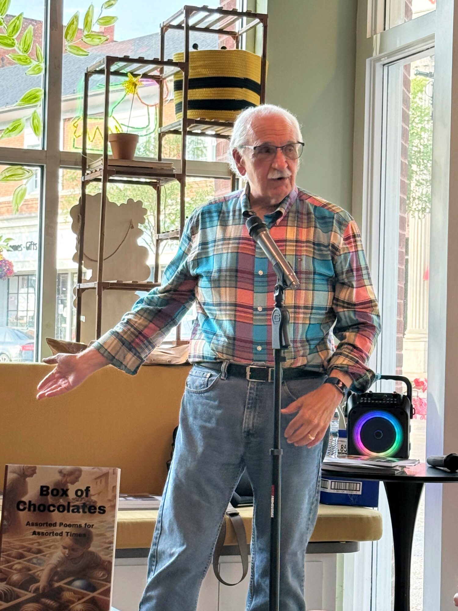 Elderly man with glasses, plaid shirt, and suspenders speaking indoors near a window, with a speaker and a book on chocolates nearby.