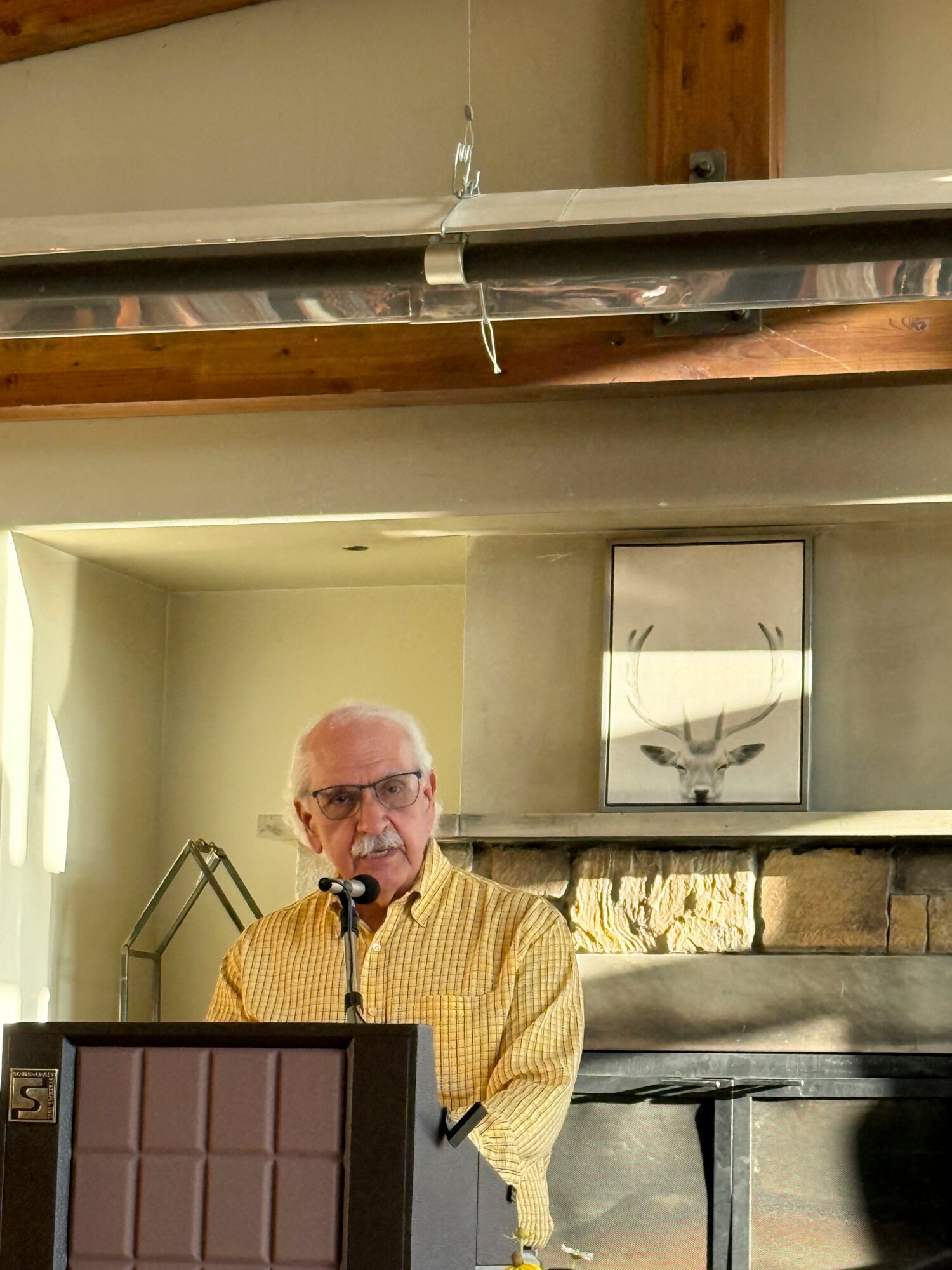 Older man with glasses and a yellow checkered shirt speaking into a microphone in a cozy room with a stone fireplace.