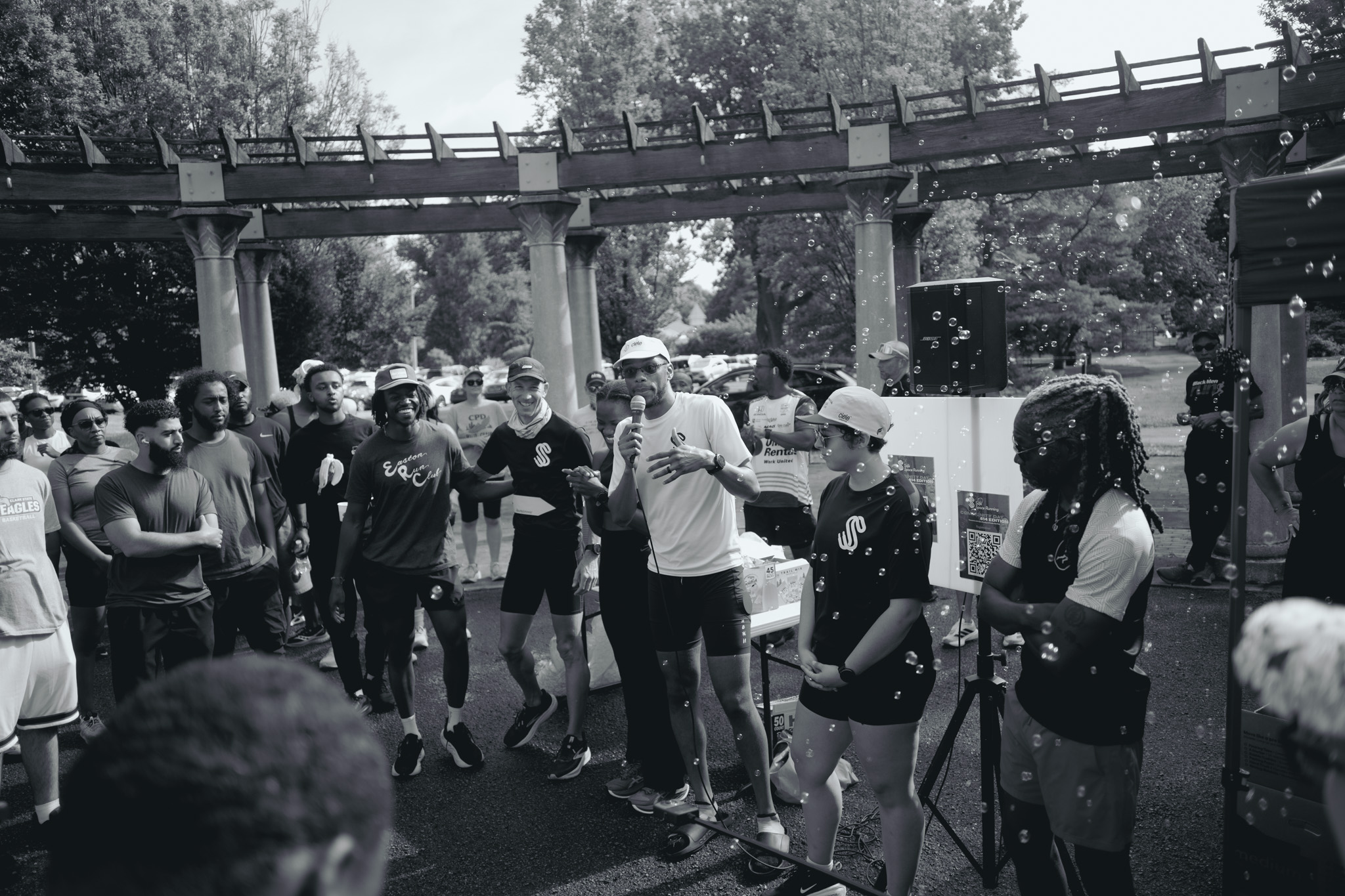 Group of people gathered outdoors near a bridge, some holding microphones, in black and white.