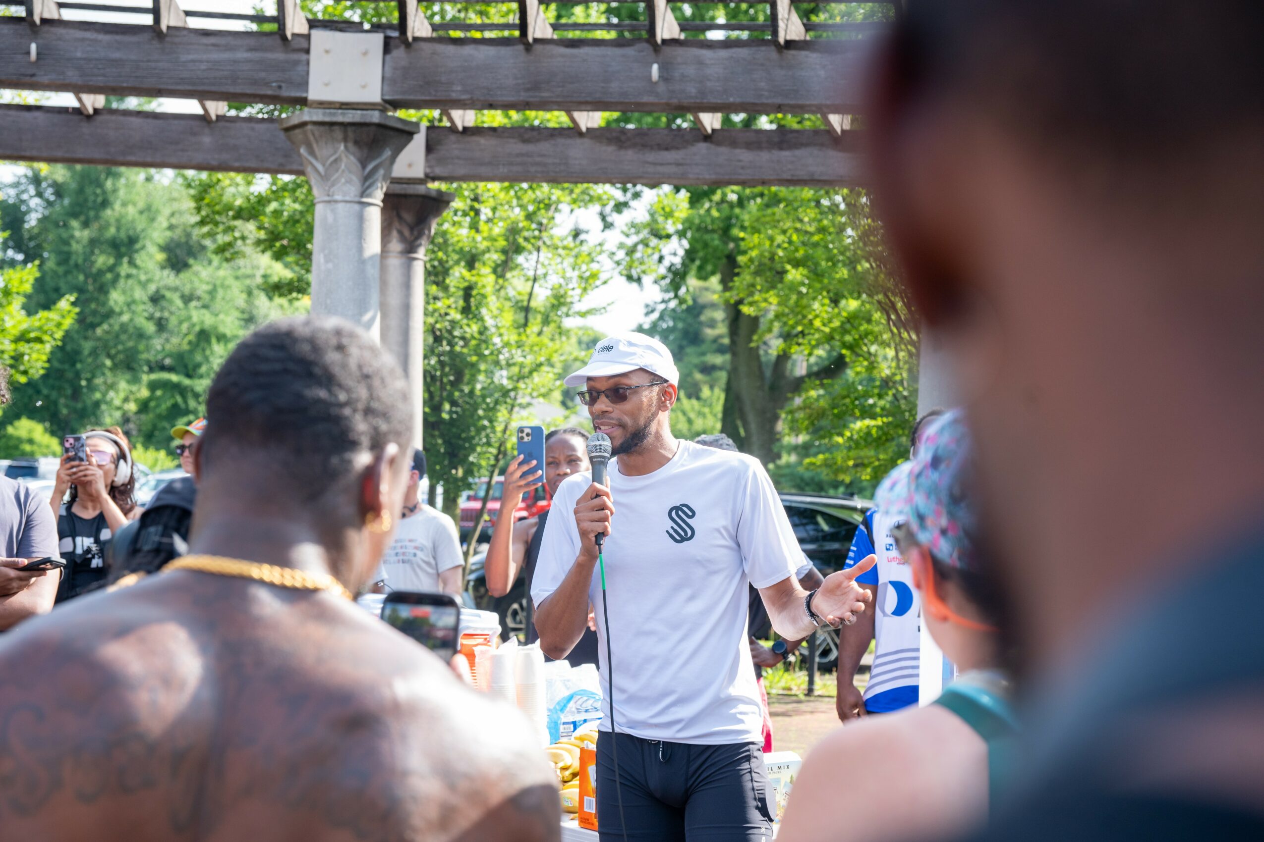 Man with a beard and sunglasses speaking into a microphone outdoors, surrounded by people and trees.
