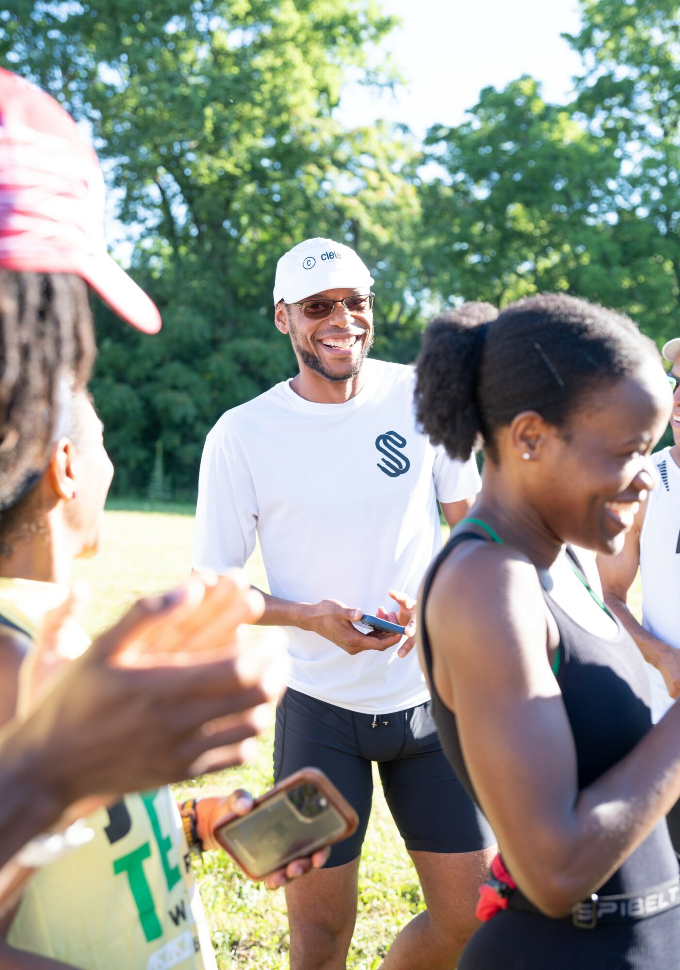 Group of people outdoors, smiling, with trees in the background, one person holding a phone, woman in foreground smiling.