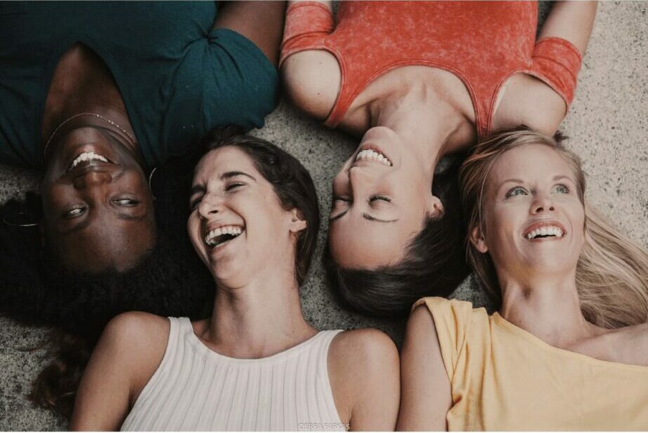 Four women lying on the floor with heads close together, smiling and looking up.