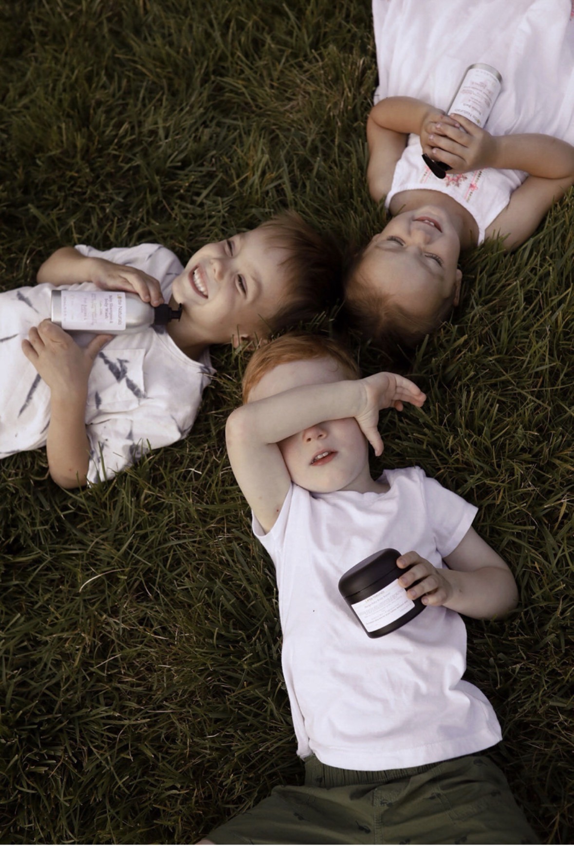 Three children lying on grass, holding drinks, one shielding eyes, one smiling, one looking up, relaxed outdoors.