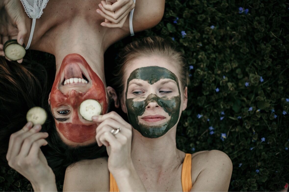 Two women lying on grass with face masks, one with red face mask and the other with green, holding fruit and smiling.