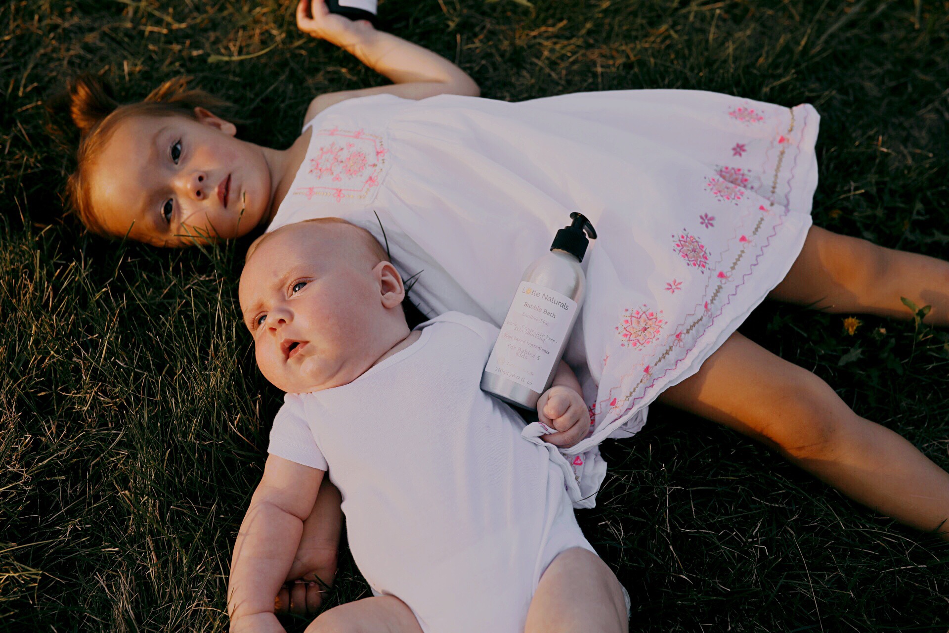Two children lying on grass, one girl in a white dress and one baby in a white onesie, with a bottle nearby.