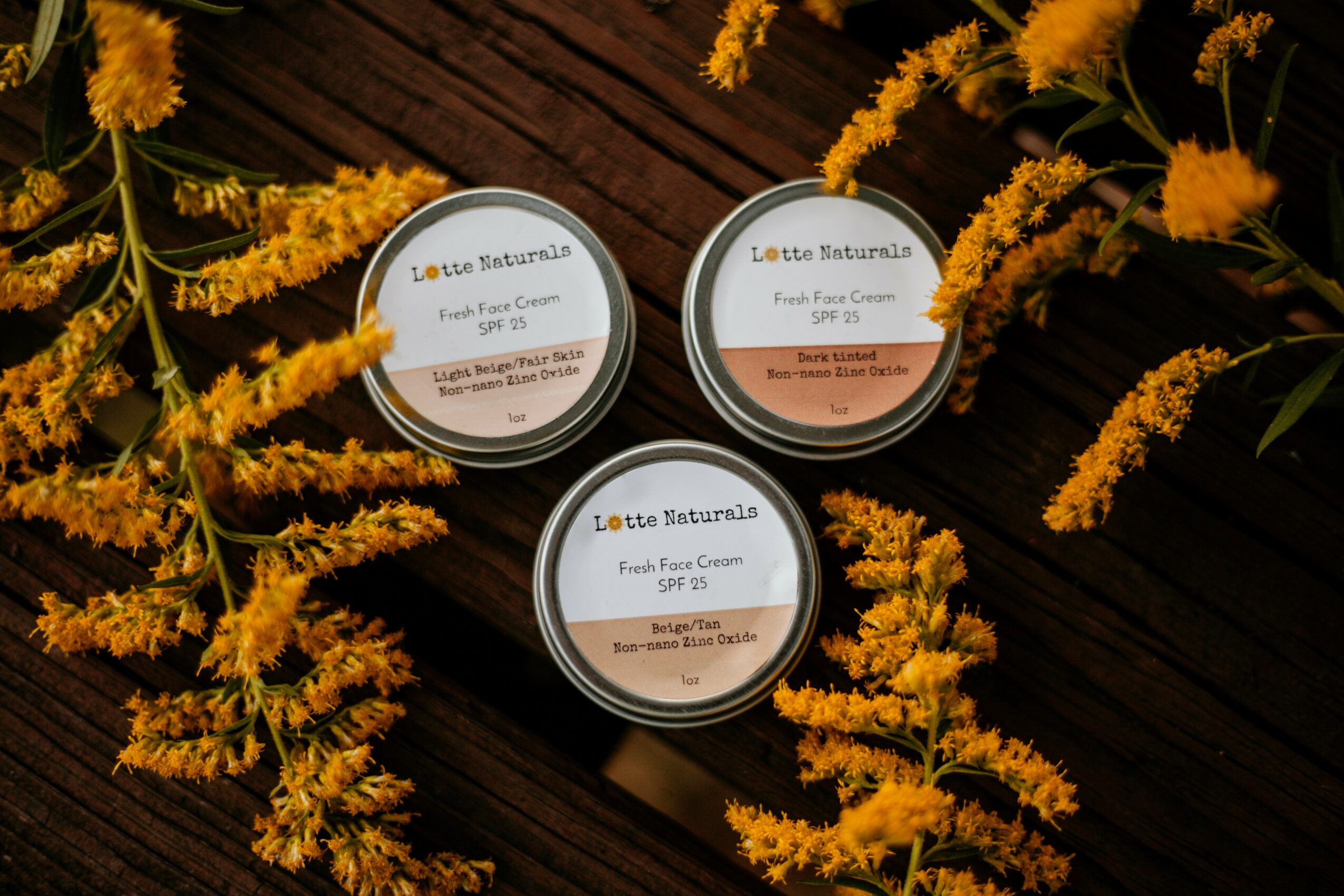 Three small containers of Lotte Asturina face cream on a dark wooden surface, surrounded by yellow flowers.
