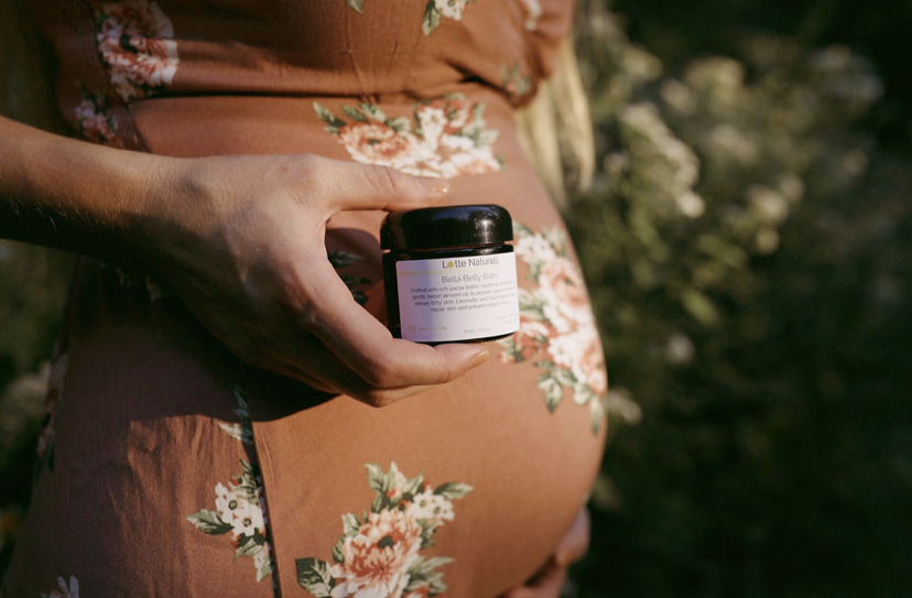 Close-up of a pregnant woman holding a small jar outdoors, wearing a floral dress, with greenery in the background.