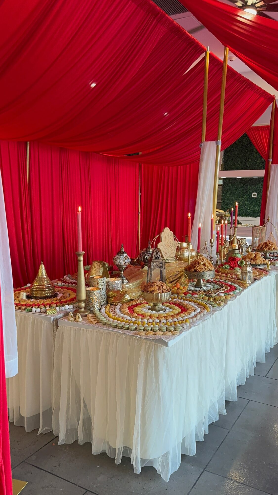 Decorated table with gold and white tableware, candles, and red drapery backdrop at an event, viewed from the side.