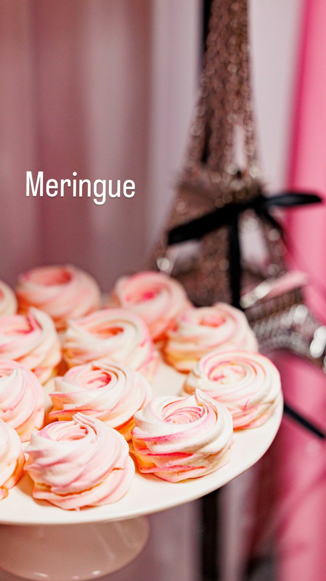 Plate of pink and white meringue cookies with a blurred background including a pink wall and decorative items.