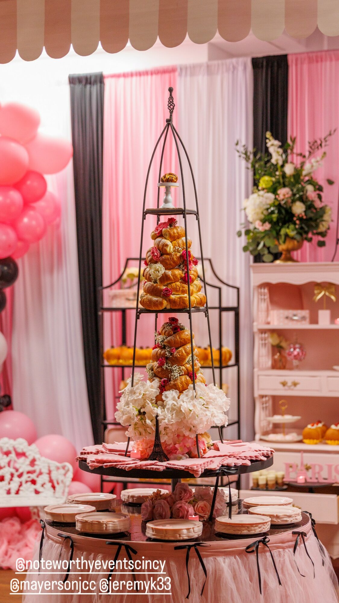 Decorative display with flowers, balloons, and a tiered stand holding desserts at a celebration, pink and black curtains in background.