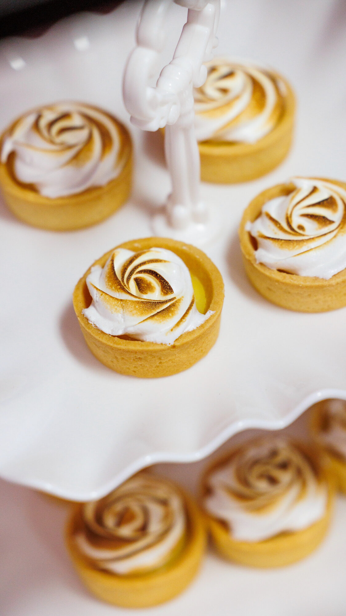 Six lemon tarts with swirled meringue topping on a white surface, with a decorative white figure in the background.