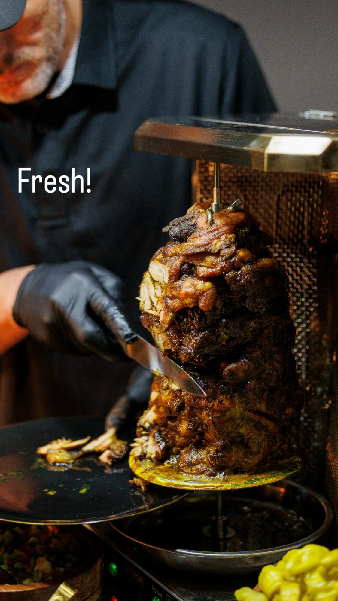 Person slicing cooked meat from a vertical rotisserie, with a sign reading 'Fresh!', black gloves, and a dark shirt.