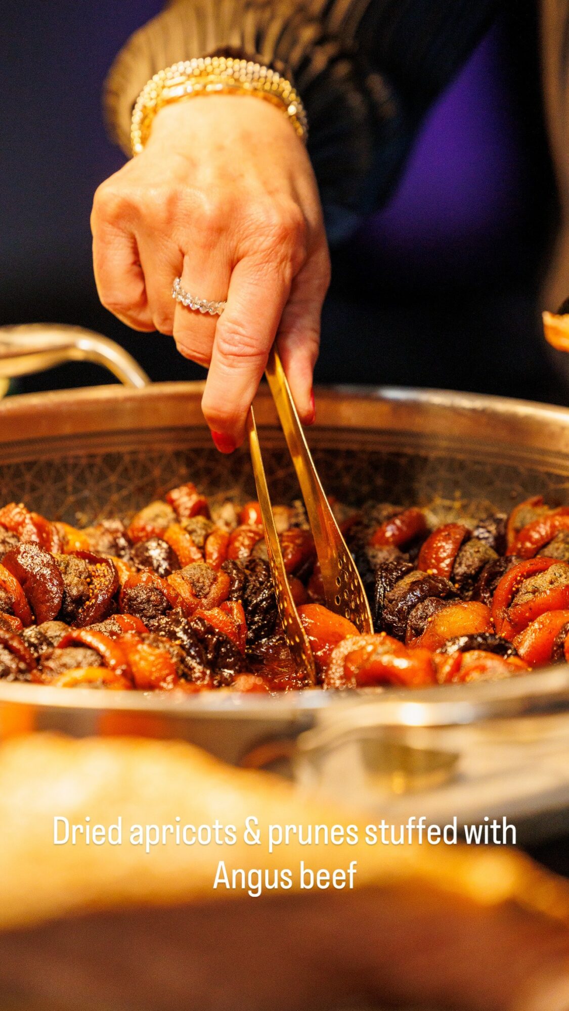Hand using tongs to serve dried apricots and prunes from a pot, with a blurred background.