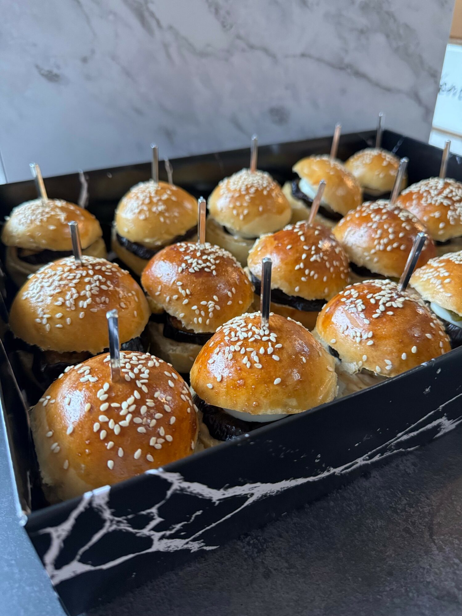 Tray of small round pastries topped with sugar, each with a small stick inserted, on a black marble-patterned surface.