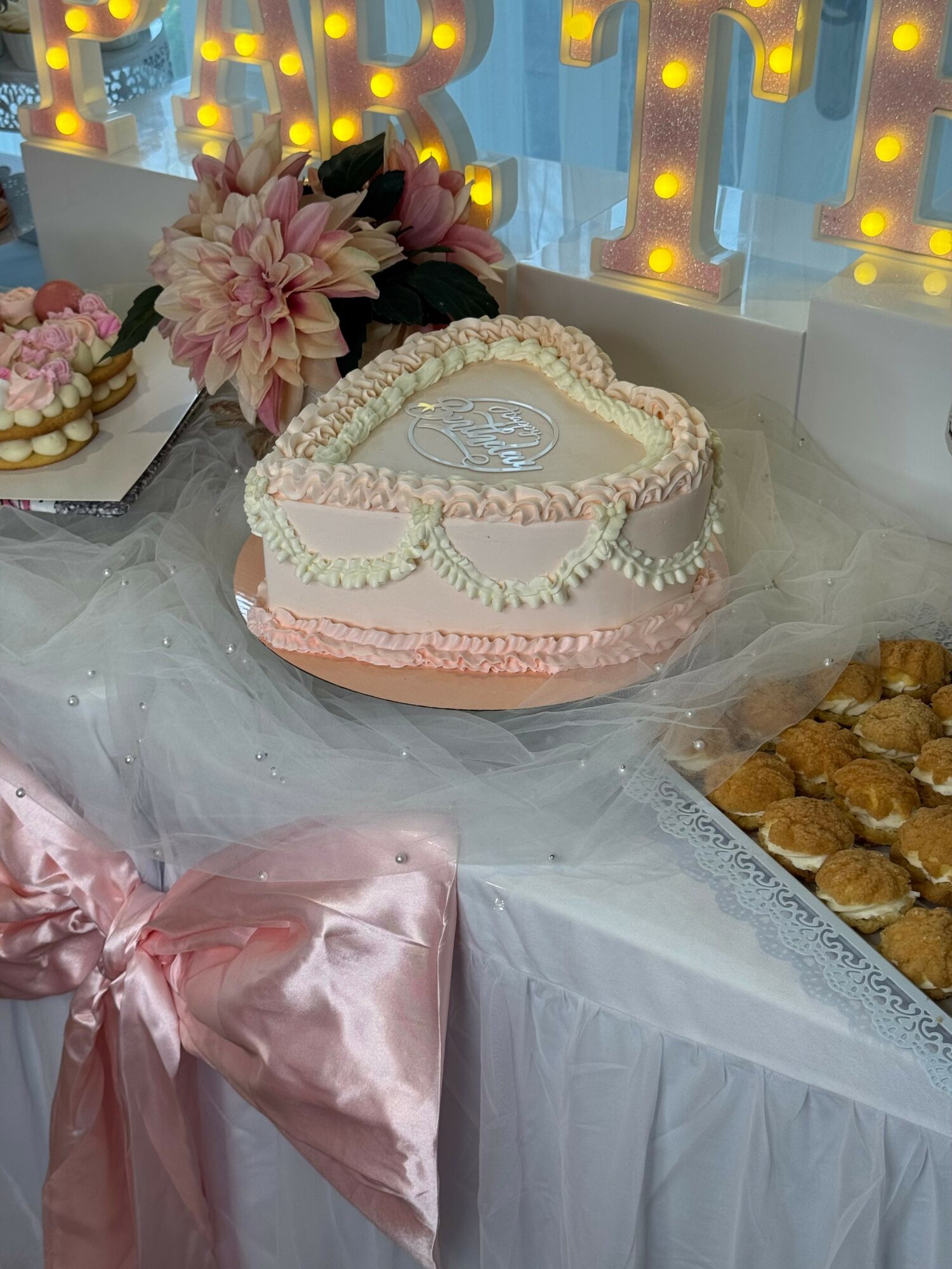 Heart-shaped cake with pink and white icing, decorated with flowers, on a table with flowers and treats, pink satin ribbon.