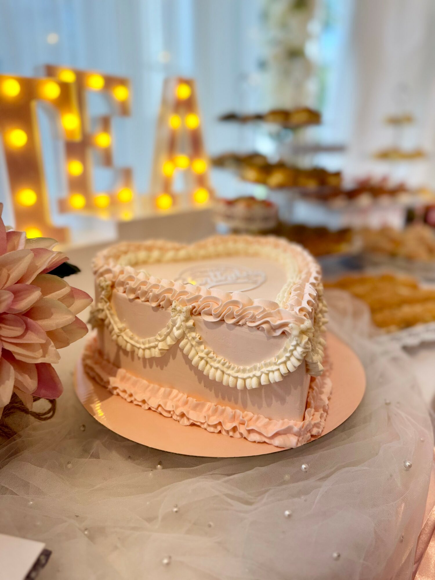 Heart-shaped cake with pink and white frosting, decorated with piped borders, on a table with a blurred dessert display in background.