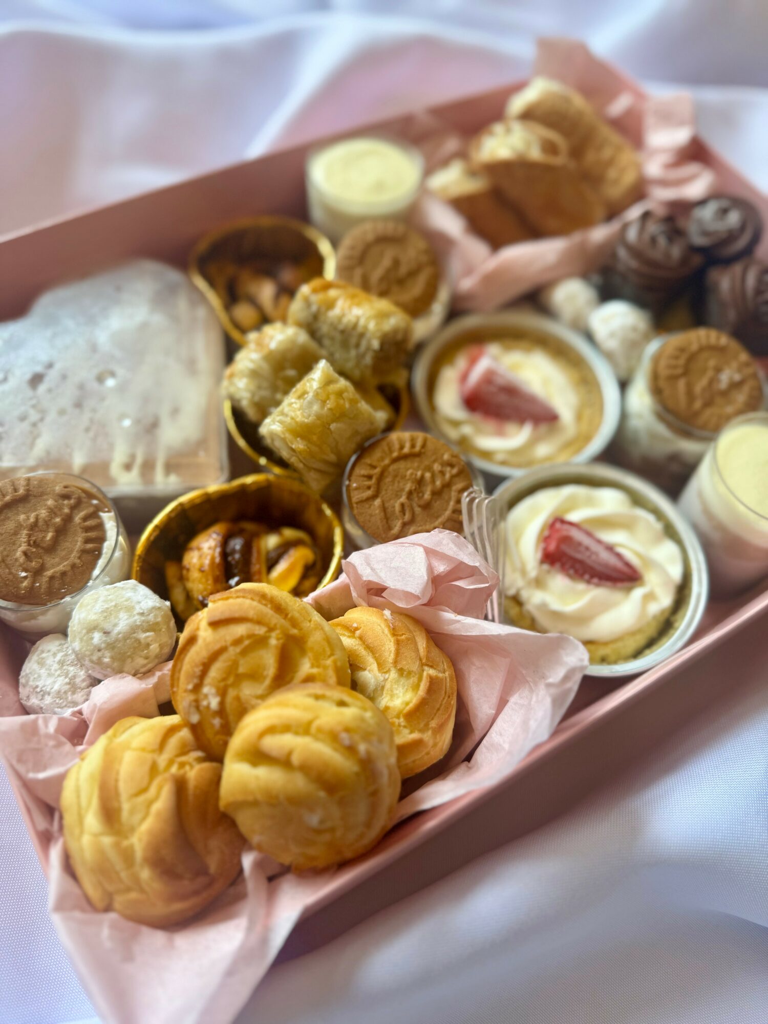 Assorted cookies, biscuits, and small desserts in a pink tray with pink paper lining, some in small cups, with a slice of bread.