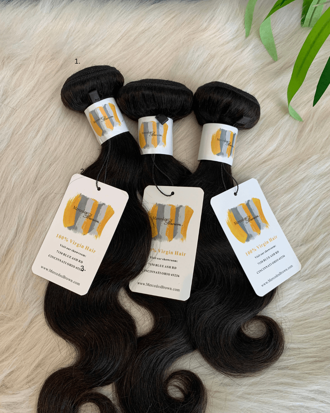 Three bundles of black hair extensions with white tags, placed on a soft, cream-colored surface with green leaves nearby.