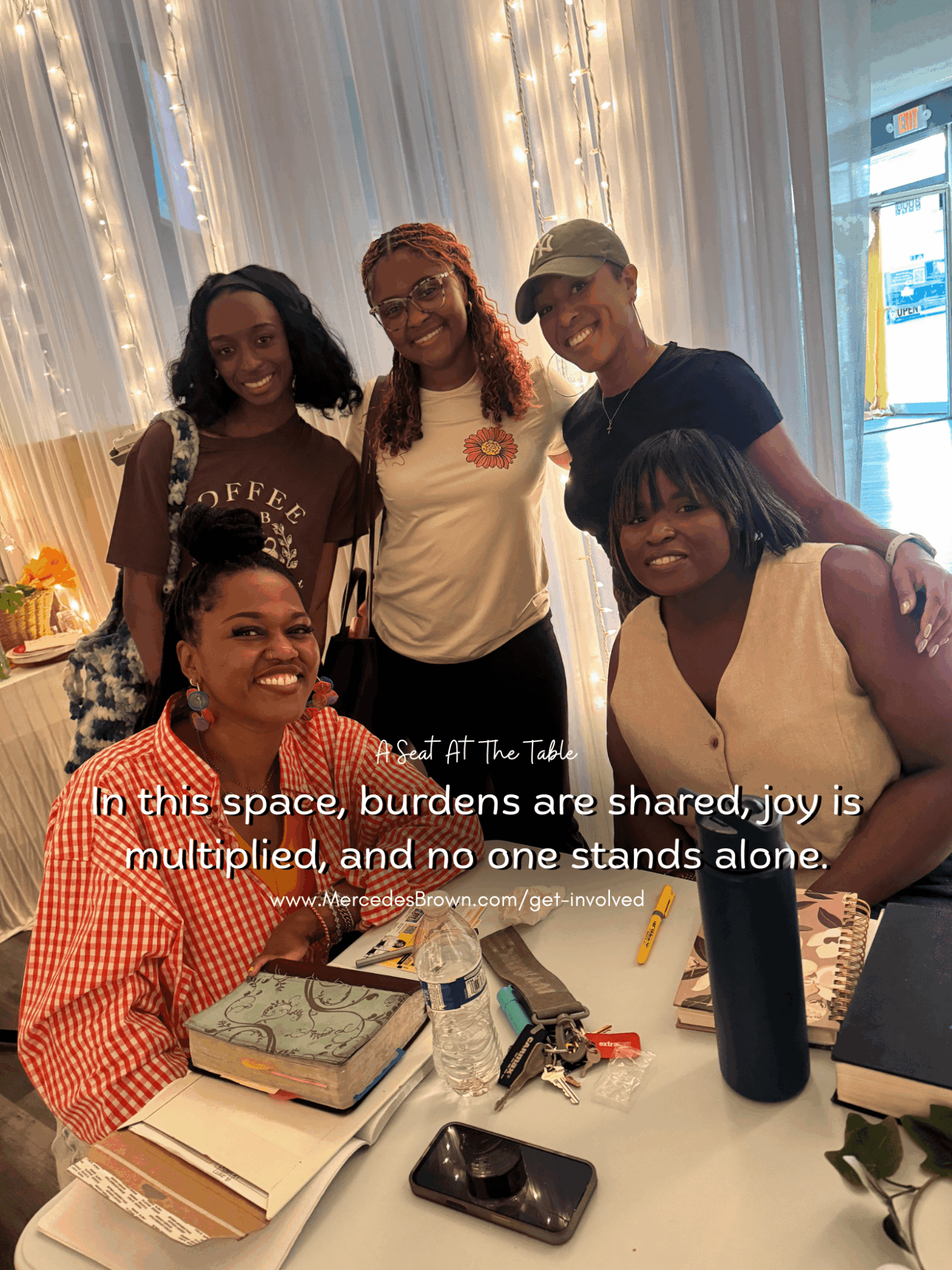 Five women gathered around a table with books and a phone, smiling indoors with curtains and string lights behind them.